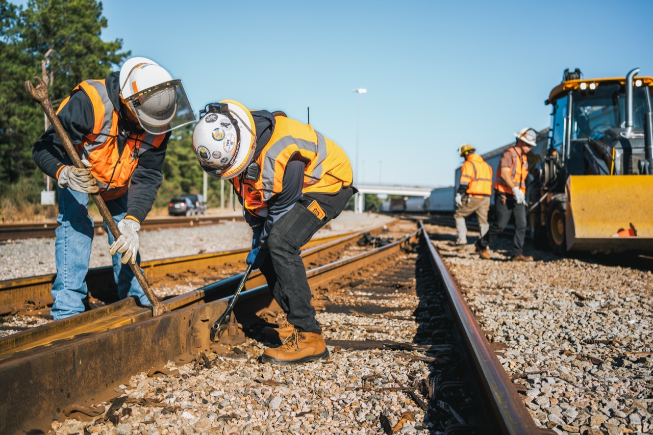 Railroad maintenance crew wearing safety gear works on tracks beside construction equipment.