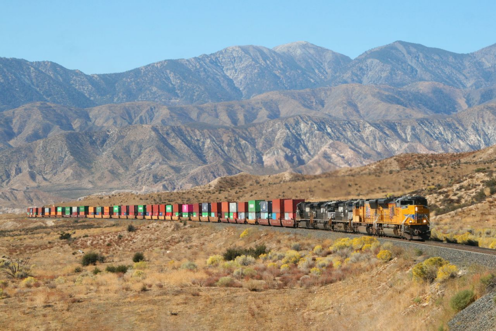 Union Pacific and Norfolk Southern locomotives lead an intermodal freight train through a mountainous desert landscape.