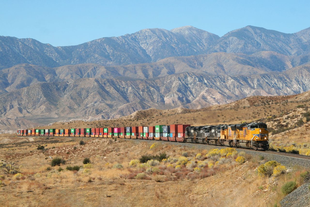 Union Pacific and Norfolk Southern locomotives lead an intermodal freight train through a mountainous desert landscape.