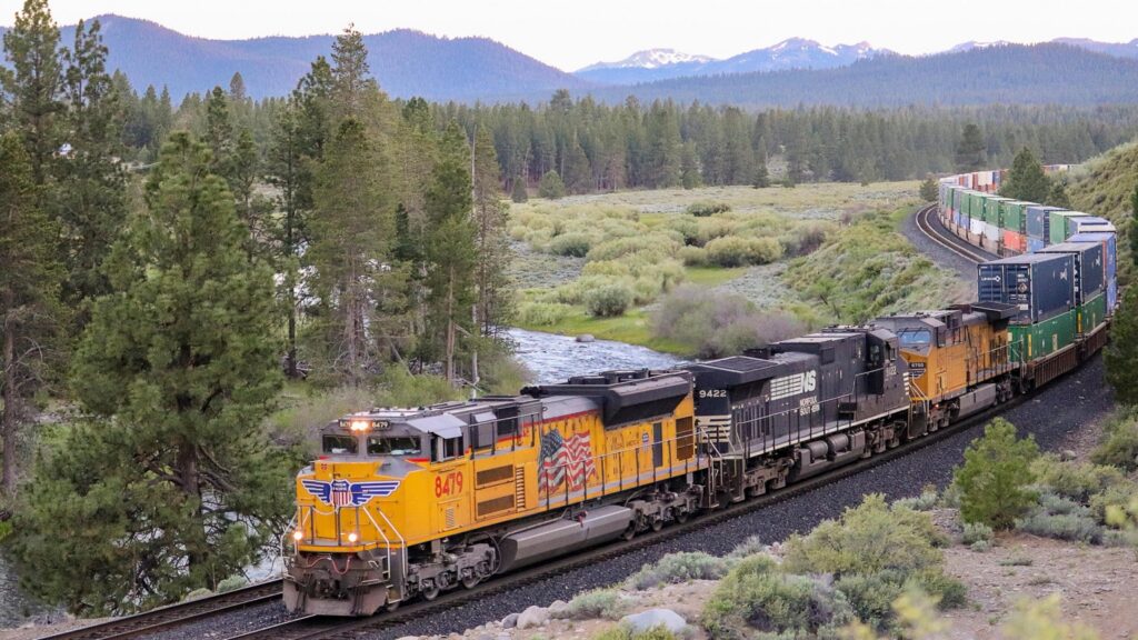 Union Pacific and Norfolk Southern locomotives pull an intermodal train beside a river through a forested mountain landscape.