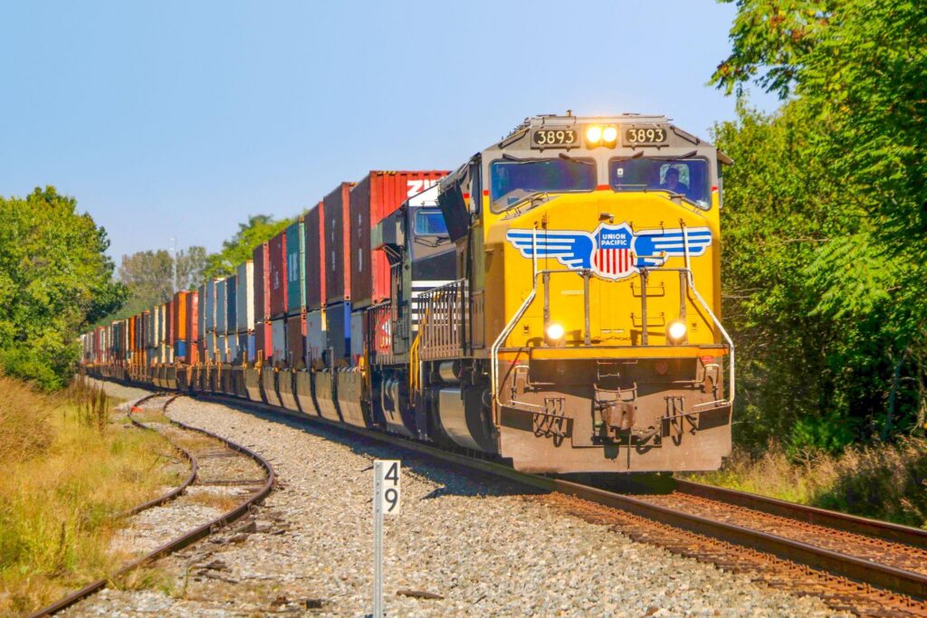 Union Pacific locomotive 3893 leads an intermodal freight train along a tree-lined rail corridor.
