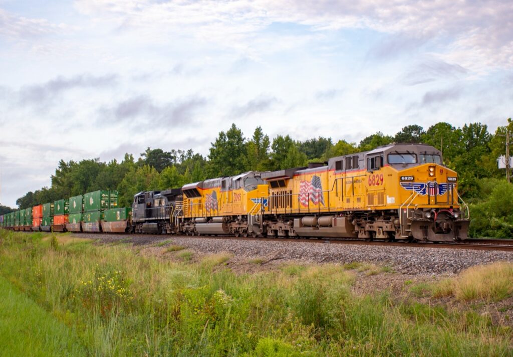 Union Pacific and Norfolk Southern locomotives lead an intermodal freight train through a green, tree-lined landscape.