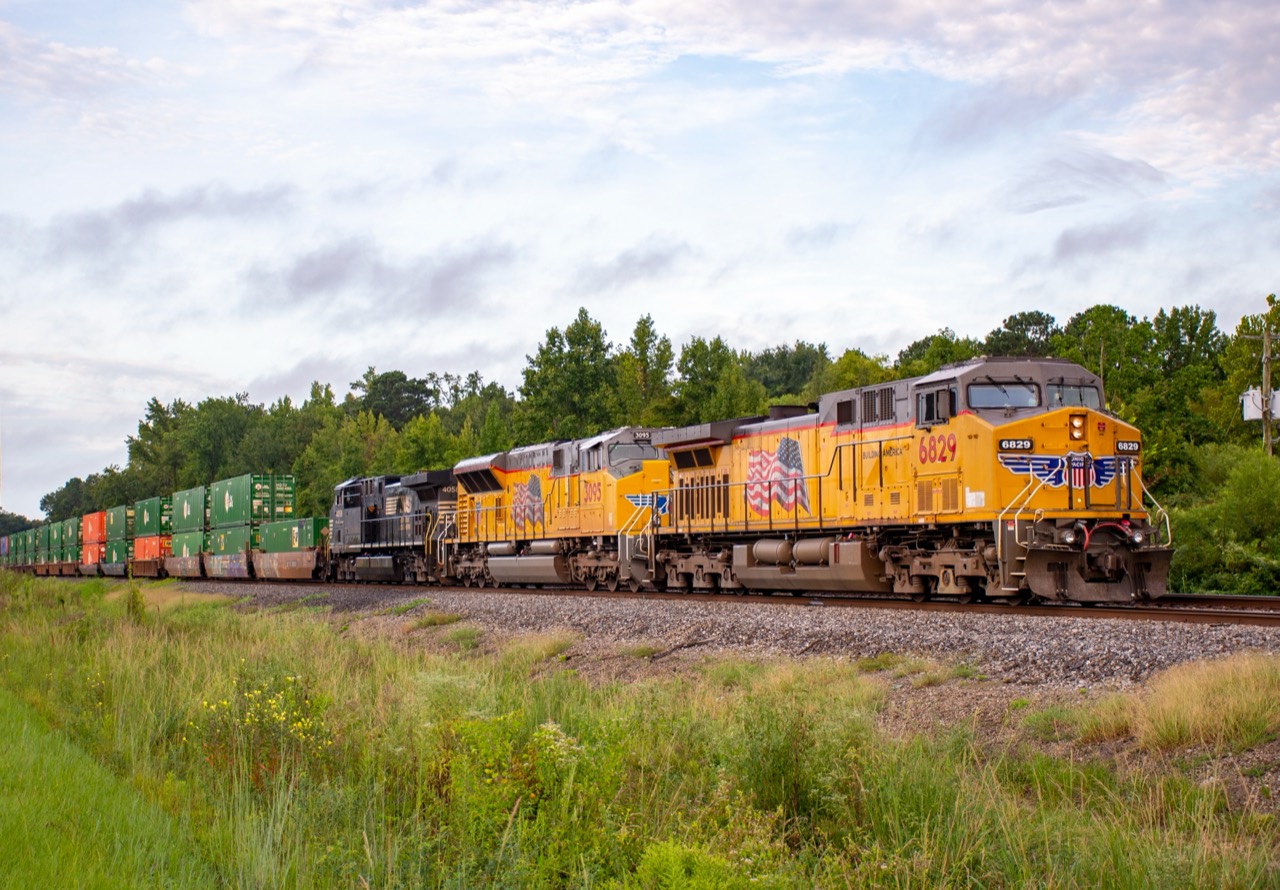 Union Pacific and Norfolk Southern locomotives lead an intermodal freight train through a green, tree-lined landscape.